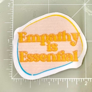 May include: A pink and white sticker with a yellow outline and blue accents. The sticker says "Empathy is Essential" in a retro font.