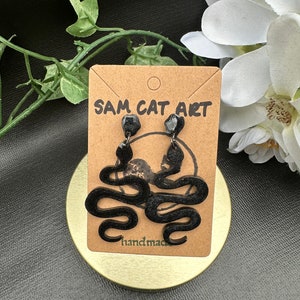 May include: Black snake-shaped earrings on a brown card with the text "SAM CAT ART" and "handmade". The earrings are displayed on a gold-colored round surface, with green and white floral accents in the background.