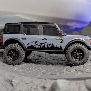 Puede incluir: Un SUV de juguete gris y negro con neumáticos negros grandes y una rueda de repuesto en la parte trasera. El SUV tiene un gráfico de montaña y árboles en el lateral. El juguete está sobre una superficie rocosa gris.