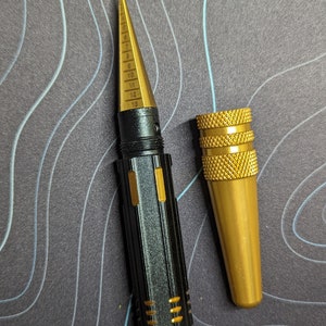 May include: A black and gold drill bit gauge with a pointed tip and a numbered scale from 5 to 69. The gauge is accompanied by a separate gold-colored drill bit holder.