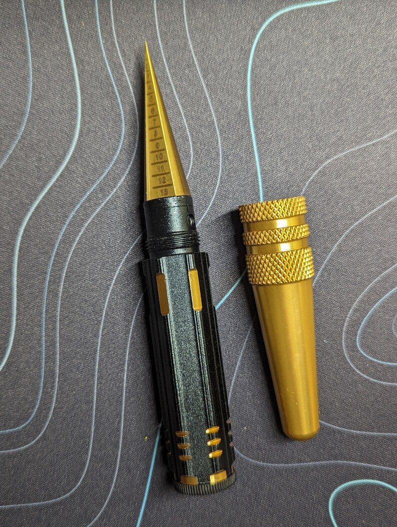 May include: A black and gold drill bit gauge with a pointed tip and a numbered scale from 5 to 69. The gauge is accompanied by a separate gold-colored drill bit holder.