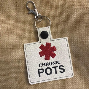 Könnte beinhalten: Weißer Leder-Schlüsselanhänger mit einem roten medizinischen Symbol und dem Text "CHRONIC POTS" in Schwarz.