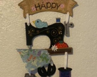 May include: A decorative sewing-themed wall hanging. It features a black sewing machine, a blue bird, fabric, scissors, a thimble, and a spool of thread. The banner above reads "Sew Happy".