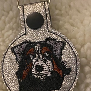May include: A silver key chain with a black button and a round, embroidered image of a Bernese Mountain Dog's face.