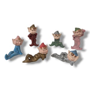 May include: A collection of seven ceramic elf figurines in various poses. Each elf has a smiling face, pointed ears, and a small hat. The elves are painted in shades of blue, red, green, brown, and silver, with pink and gold accents. They are arranged on a white background.