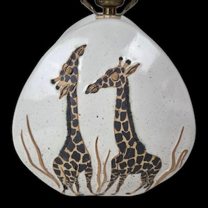 May include: A teardrop-shaped white ceramic plaque with a speckled finish featuring two raised, textured giraffes with brown and black spot patterns. The giraffes are positioned among thin, light brown sculpted grass elements on the surface.