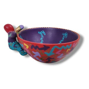 May include: A small, hand-painted ceramic bowl with a purple interior and a red exterior. The bowl features a whimsical design with colourful swirls and abstract shapes. A small, colourful figure is attached to the side of the bowl, acting as a handle.