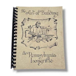 May include: A spiral-bound book titled The Art of Building the Pennsylvania Longrifle. The cover is tan with a black ink illustration of a person working on a rifle at a workbench surrounded by various woodworking tools on a scroll background.
