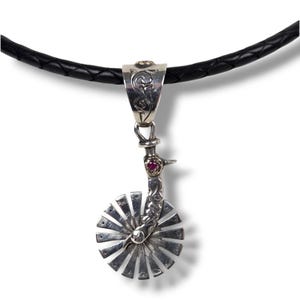 May include: A silver pendant necklace featuring a spur design. The pendant has a circular, sunburst-like element and a decorative arm with a small red gemstone. The necklace has a black leather cord.