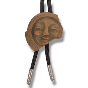 May include: A bronze-colored bolo tie featuring a smiling face design. The pendant is attached to a black braided cord with silver-colored metal tips. The smiling face has closed eyes and a slightly open mouth.