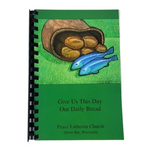 May include: A green book with a spiral binding. The cover features an illustration of a basket of bread and two blue fish. The title of the book is "Give Us This Day Our Daily Bread". The book is from Peace Lutheran Church in Green Bay, Wisconsin.