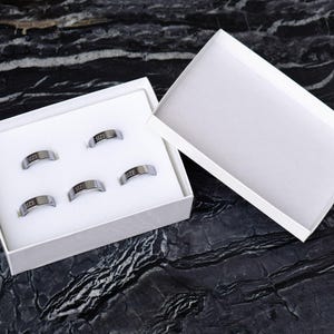 May include: A set of five silver-toned metal rings arranged in a white rectangular box with a matching lid. Each ring features the word SIZE engraved in black lettering on the outer surface. The rings are held in place by a white foam insert.