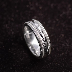 May include: A silver-toned metal ring featuring a polished finish and a central band decorated with a repeating etched leaf or chevron pattern. The design is bordered by two thin, raised beaded edges that run along the circumference of the band.