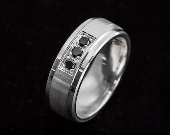 May include: A silver-toned metal ring featuring a brushed finish on the outer band and a polished centre channel. The ring is set with three small, round, black stones arranged in a vertical row within a rectangular recessed setting.