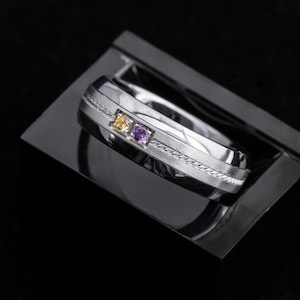 May include: A silver-toned metal ring featuring a brushed finish and a central braided texture band. The ring is set with two small round-cut gemstones, one yellow and one purple, positioned side-by-side in a square-shaped metal setting.