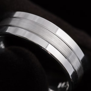 Könnte beinhalten: Ein silberfarbener Metallring mit einem breiten, gebürsteten Mittelband, das von zwei polierten, reflektierenden Kanten flankiert wird. Das Design zeichnet sich durch seine klare metallische Oberfläche und den Kontrast zwischen der matten Textur und den glatten Rändern aus.