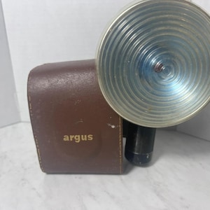 May include: A vintage Argus camera flash unit featuring a brown leather-textured casing with the word argus printed in gold lettering. The device includes a large, circular, silver-toned metallic reflector with concentric circular ridges.