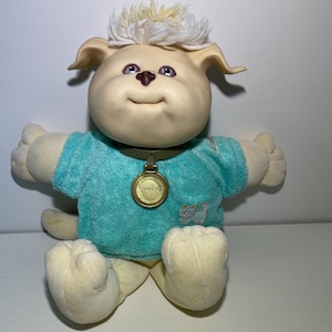 May include: A vintage plush dog toy with a cream-coloured face, ears, and paws. It has a light blue shirt and a fluffy, cream-coloured hairpiece. The dog has a gold-coloured tag around its neck and a small cat graphic on the shirt.