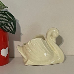 May include: A cream ceramic swan planter with a curved neck and detailed wing feathers. The planter has a small opening for plants. A red planter with a white heart is visible on the left.