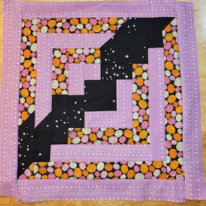 May include: A purple and black quilt with a geometric pattern of squares and triangles. The quilt is made of fabric with a pattern of orange, pink, and white pumpkins.