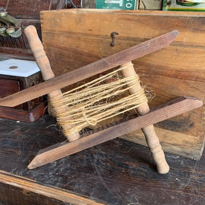 May include: A wooden twine holder with a spool of twine wound around it. The twine holder is made of two pieces of wood that are joined together at a right angle. The twine is wrapped around the holder and is held in place by a wooden peg.