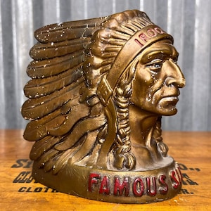 May include: A bronze-colored bust of a Native American chief with a feathered headdress. The word "IROQUOIS" is visible on the headdress, and the word "FAMOUS" is in red lettering on the base. The bust has a weathered appearance.