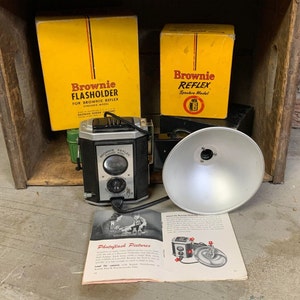 May include: A vintage Brownie Reflex Synchro Model camera with a flash attachment and instruction manual. The camera is black with chrome accents and has a flash holder attached to the top. The instruction manual is open to a page about photoflash pictures.
