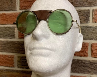 May include: Vintage green tinted round safety glasses with a brown leather brow guard. The glasses are on a white mannequin head.