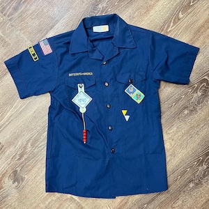 May include: A navy blue Boy Scouts of America uniform shirt with short sleeves. The shirt has a pocket on the left chest with the words "Boy Scouts of America" embroidered in yellow. There is a small American flag patch on the right sleeve and a variety of other patches on the front of the shirt.