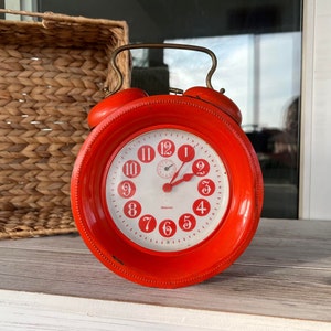May include: A bright red vintage alarm clock with white face and black hands. The clock has a bell on each side and the numbers 1-12 are in red.