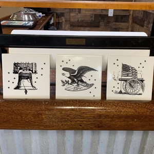 May include: Three white ceramic tiles with black and white patriotic images. The first tile features a bell with stars, the second tile features an eagle with stars, and the third tile features a cannon with an American flag and stars.