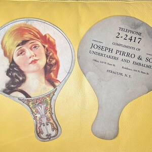 May include: A vintage hand fan with a woman's face on one side and an advertisement for Joseph Pirro & Son Undertakers and Embalmers on the other side. The fan is made of paper and has a decorative handle.