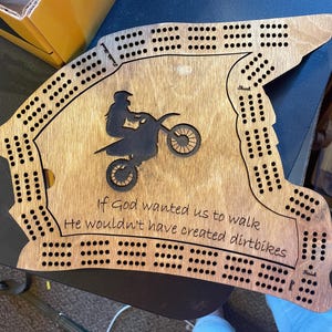 May include: Wooden cribbage board with a motorcycle silhouette and the quote, "If God wanted us to walk, He wouldn't have created dirtbikes." The board has holes for pegs and is shaped like a motorcycle helmet.