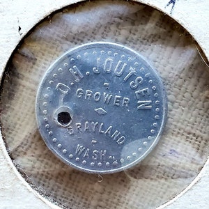 May include: A silver token with the text "D.H. JOUTSEN GROWER GRAYLAND WASH."  The token is in a clear plastic display case with a white cardboard backing. The cardboard backing has the text "Good" written on it.