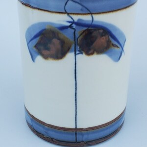 May include: A white ceramic utensil holder with a blue and brown abstract design. The holder has a cylindrical shape and a smooth, glazed finish.