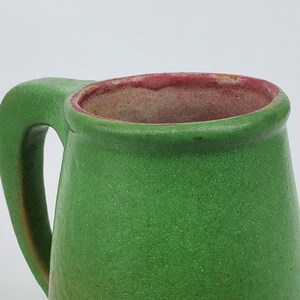 May include: A green ceramic mug with a large handle. The mug has a textured surface and a reddish-pink rim. The interior of the mug is a light brown color. The mug is a simple, yet elegant design.