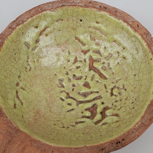 May include: A small, green ceramic bowl with a brown handle. The bowl has a textured surface and a unique design.