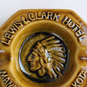 May include: A brown ceramic ashtray with a Native American chief design and the text "Lewis & Clark Hotel Mandan North Dakota".