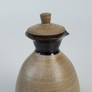 May include: A ceramic pitcher with a lid, featuring a light brown glaze and a dark brown band around the neck. The pitcher has a rounded body and a slightly flared lip.