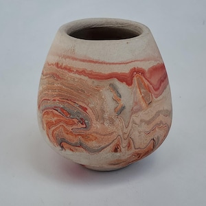 May include: A small, egg-shaped ceramic vase with a wide opening. The vase has a marbled design with swirls of red, orange, grey, and cream colours. The interior is a darker brown.