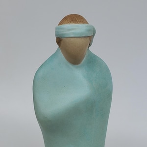 May include: A ceramic figurine of a person wearing a light blue robe and a light blue headband. The figurine is standing on a white surface.
