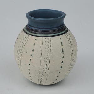 May include: A handcrafted ceramic vase with a round body and a blue rim. The off-white surface is decorated with vertical lines of dotted and triangular patterns. The vase is approximately 6 inches tall.