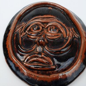 May include: A round ceramic wall hanging with a brown and black glaze. The hanging features a stylized face with exaggerated features.