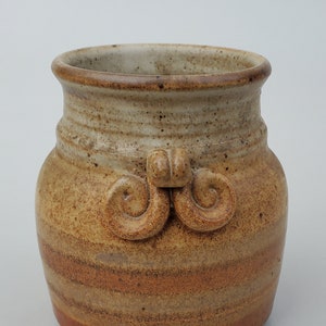 May include: A small, brown and tan ceramic vase with a decorative design. The vase has a rounded body and a wide mouth.