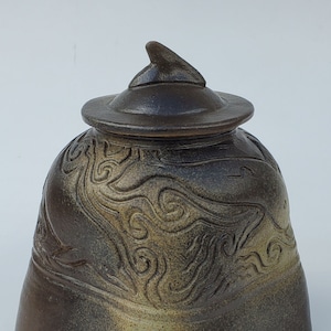 May include: A brown ceramic jar with a lid and a swirling pattern. The jar has a pointed lid and a wide base.