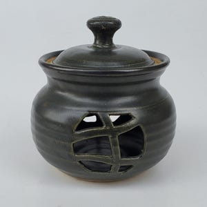 May include: A dark green ceramic garlic keeper with a lid and a knob handle. The jar has a round shape with a decorative cut-out design on the front. The lid fits snugly on top, and the piece has a rustic, handmade appearance.