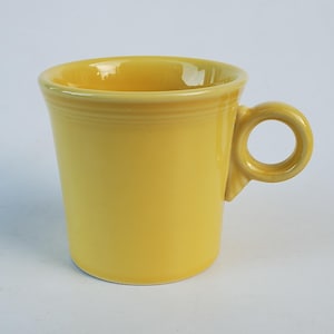 May include: A bright yellow ceramic mug with a rounded handle. The mug has a slightly flared shape and a smooth, glossy finish. The handle is a circular loop attached to the side of the mug.