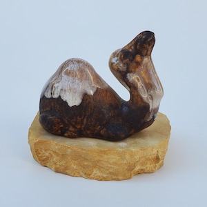 May include: A ceramic camel figurine with brown and white glaze, sitting on a small, light brown stone base.