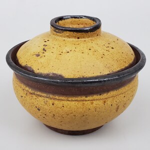 May include: A yellow ceramic bowl with a lid, featuring a brown and black rim. The bowl has a speckled glaze and a rounded shape.