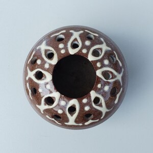 May include: A brown ceramic incense holder with a circular design. The holder features a central opening and is adorned with white, eye-shaped patterns and small perforations. The design is symmetrical and the object is set against a white background.
